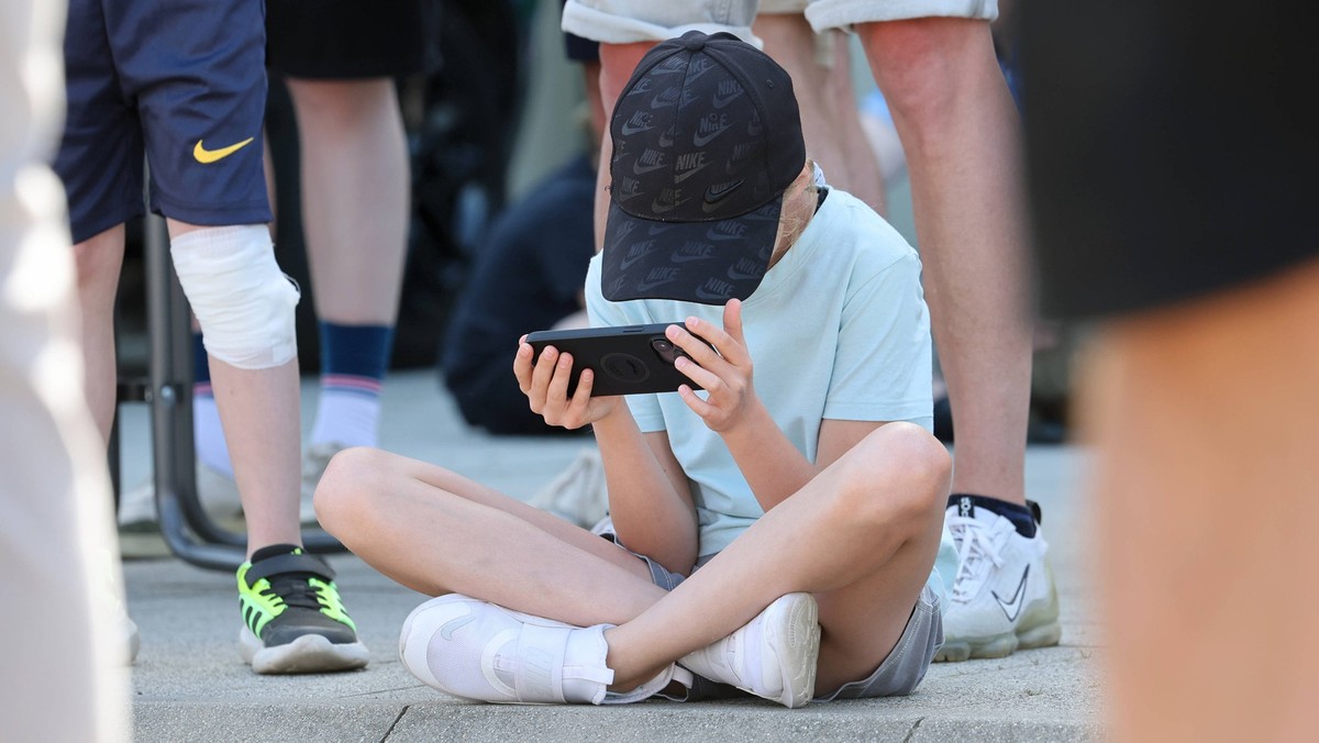 30.05.2026, Leipzig, Sachsen, GER - Maedchen sitzt im Schneidersitz zwischen Menschen auf dem Asphalt und schaut auf ihr Smartphone. 10-15 Jahre, Alltag, aussen, Aussenaufnahme, Basecap, deutsch, Deut ...