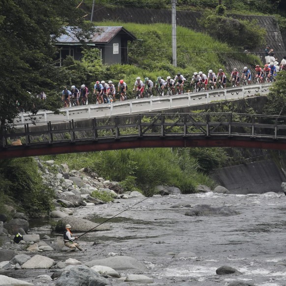A man fishes in the Doshi River as cyclists compete during a road cycling test event for the Tokyo 2020 Olympic Games, Sunday, July 21, 2019, in Yamanashi Prefecture, southwest of Tokyo. (AP Photo/Jae ...