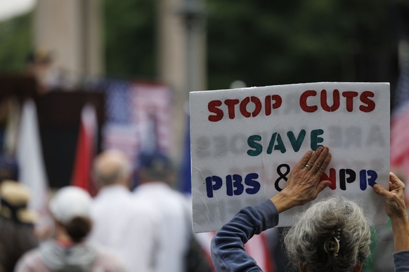 epa12161268 A demonstrator displays a sign that reads 'Stop Cuts Save PBS &amp; NPR,' while taking part in a 'No Kings on D-Day,' rally on the Boston Common, in remembrance of past ...