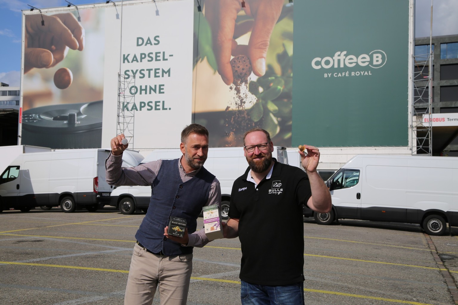 Markenbotschafter Marco Fritsche und Bulls-Coffee-Gründer Ole Bull zeigen ihre Kaffeekapseln vor einer Migros-Werbung. 