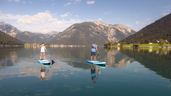 Mit einem großen, breiten Surfbrett und einem Paddel gleitet man entspannt über das Wasser des Achensees. /// Explore Lake Achensee at a leisurely pace as you stand on a large surfboard and use a padd ...