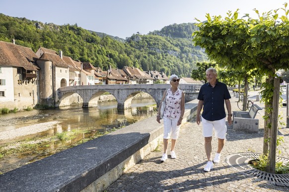 Zwei Personen spazieren am Flussufer von Saint-Ursanne am Doubs. Im Hintergrund die malerische Altstadt von Saint-Ursanne.