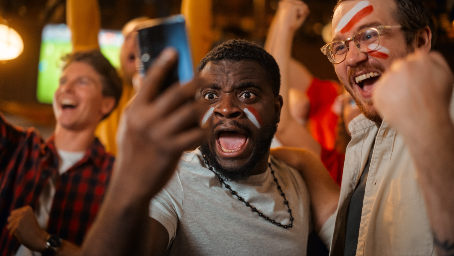 Portrait von zwei begeisterten Freunden, die ein Smartphone halten, feiern einen Sportwettkampf in ihrem Lieblingsfußballteam. Lebhafte erfolgreiche Emotionen, wenn das Fußballteam ein Gewinnziel erre ...