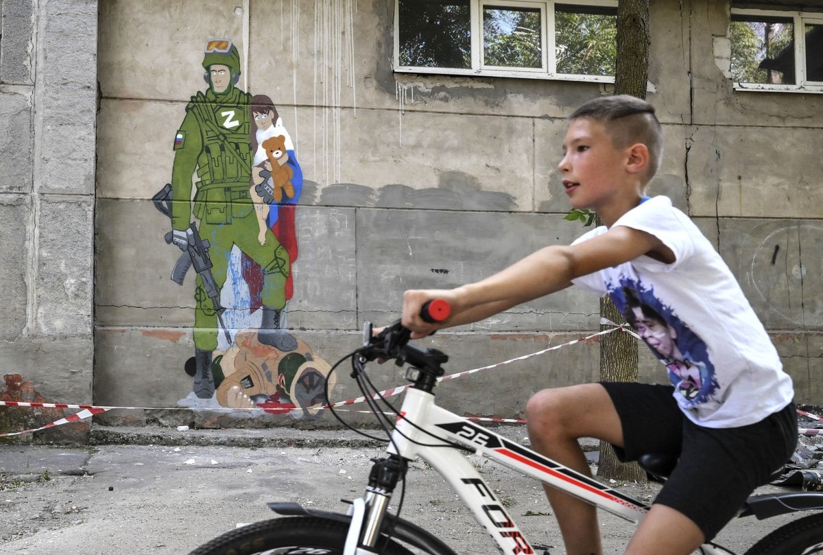 Ein ukrainischer Junge fährt in Mariupol an einem Graffiti auf einer Schulwand vorbei, das einen russischen Soldaten zeigt, der ein Kind in russischer Flagge mit Teddybär umarmt.
