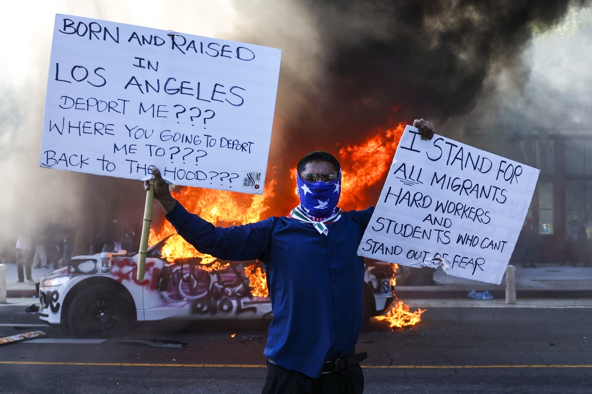 epa12165564 An activist holds signs as a car burns during protests sparked by immigration raids in Los Angeles, California, USA, 08 June 2025. US President Donald Trump has deployed 2,000 National Gua ...