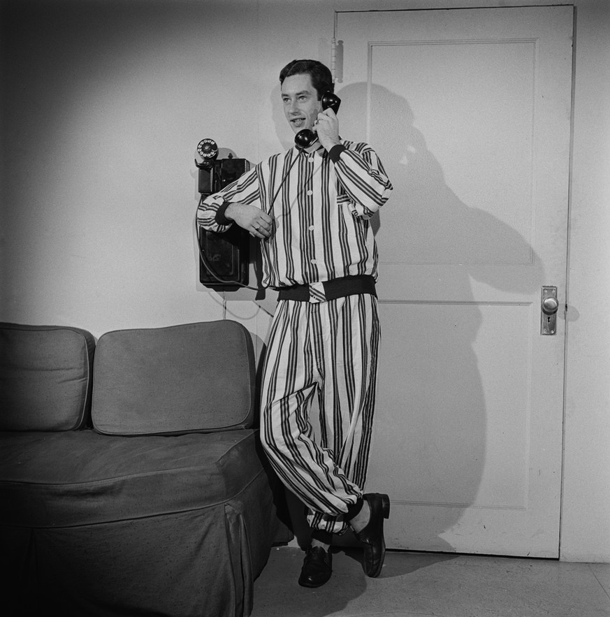A man wearing a striped outfit using a wall-mounted telephone stands beside a sofa, United States, circa 1950. The original caption read 'Pajama Fashion 1950'. (Photo by Fuchs/Graphic House/ ...