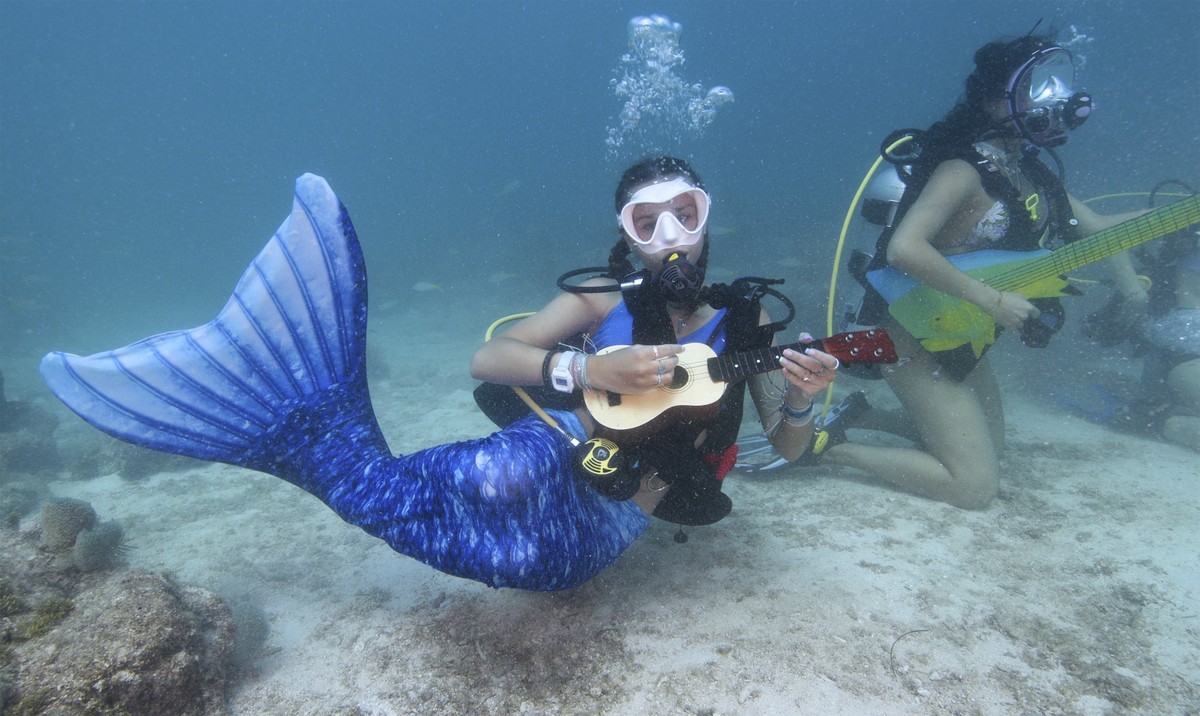 In this photo provided by the Florida Keys News Bureau, Tamara Bredova, left, costumed as a mermaid, pretends to play a ukulele underwater, Saturday, July 8, 2023, while Kelly Angel, right, strokes a  ...