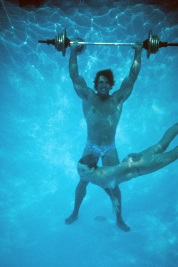 LOS ANGELES - CIRCA 1977: Austrian Bodybuilder Arnold Schwarzenegger lifts a barbell at the bottom of a pool as a topless girl swims by circa 1977 in Los Angeles, California. (Photo by Michael Ochs Ar ...