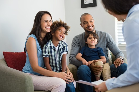 Fröhliche junge Familie sitzt auf dem Sofa und spricht mit Familienberater. Lächelnde Eltern mit adoptierten Kindern diskutieren mit Berater. Multi-ethnische Familie treffen Finanzagenten.