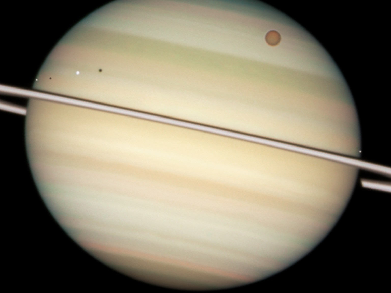 This close-up view of Saturn&#039;s disc captures the transit of several moons across the face of the gas giant planet. The giant orange moon Titan - larger than the planet Mercury - can be seen at up ...