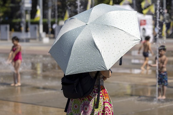 KEYPIX - A woman under umbrella for protection form the sun passes in front of children who cool off in a water-jet fountain on the Place des Nations, during a heatwave alert, in Geneva, Switzerland,  ...