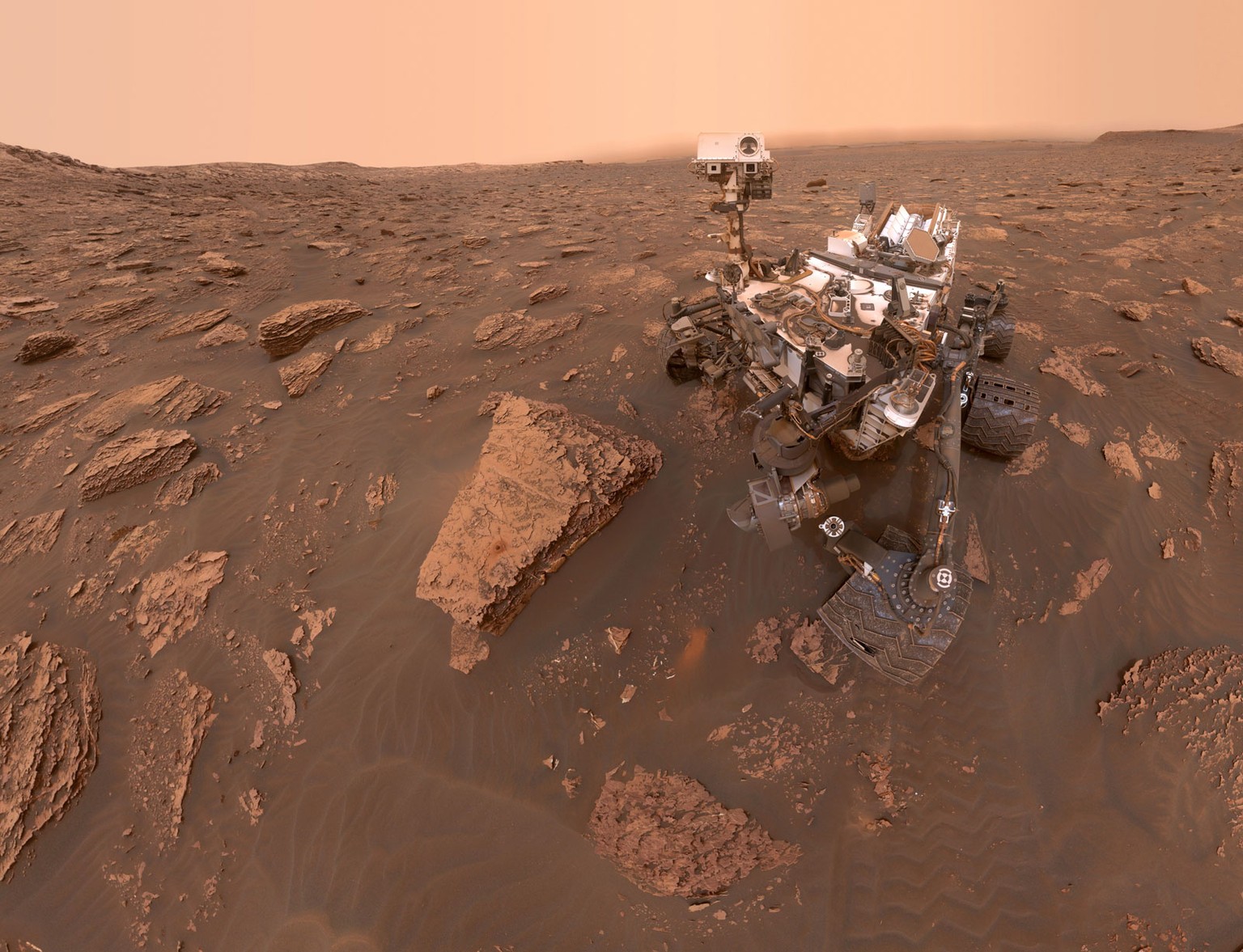 Dieses Selfie des Mars-Rovers Curiosity entstand am 15. Juni 2018. Der aufziehende Sandsturm ist am verdüsterten Horizont zu sehen. Da Curiosity – der Rover ist seit 2011 auf dem Mars – seine Energie im Gegensatz zu Spirit und Opportunity aus einer Plutoniumbatterie bezieht, kann der Staub seine Energieversorgung nicht beeinträchtigen. Das Selfie besteht übrigens aus einem Mosaik verschiedener Aufnahmen, deshalb ist der Kamera-Arm nicht zu sehen.&nbsp; &nbsp;