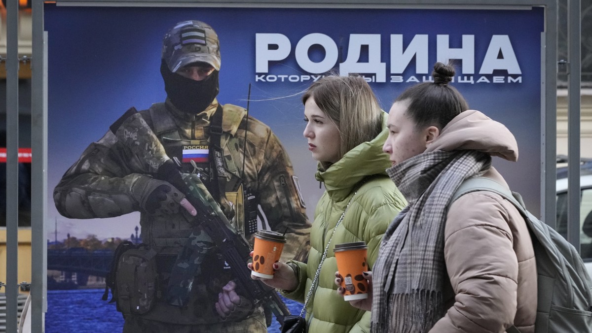 FILE - Girls walk past a stand with an image of a Russian serviceman and words 'The Motherland we defend' at a street exhibition of military photos in St. Petersburg, Russia, Feb. 9, 2023. U ...