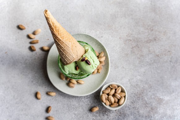 A scoop of pistachio ice cream on a plate, topped with a waffle cone and surrounded by scattered pistachios. A bowl of pistachios complements the arrangement on a textured grey surface. Model Released ...