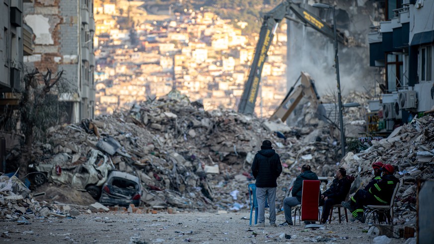 epa10478168 People sit in front of collapsed buildings after a powerful earthquake, in Hatay, Turkey, 19 February 2023. More than 46,000 people have died and thousands more are injured after two major ...