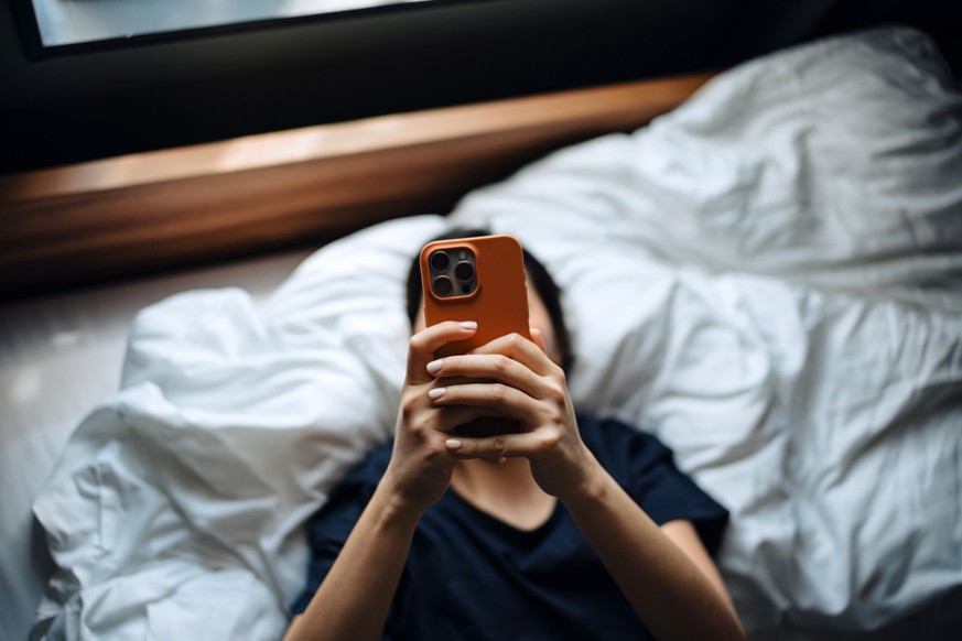 High angle shot of young Asian woman texting on her smartphone while lying on bed relaxing at cozy home. Chatting online with friends. Social media. Youth culture. Lifestyle and technology
