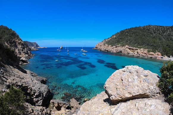 A tranquil scene of sailboats at sea, visible from the rocky coast in the foreground, Ibiza Model Released Property Released xkwx beach blue boat calm clouds coast cruise dock horizon ibiza landscape  ...