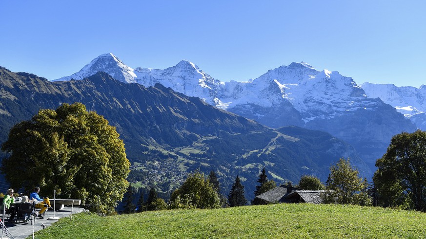 Personen geniessen auf einer Terasse das schoene Herbstwetter mit Blick auf das Dreigestirn Eiger, Moench und Jungfrau, von links, am Sonntag, 16. Oktober, in Sulwald ob dem Lauterbrunnental. (KEYSTON ...