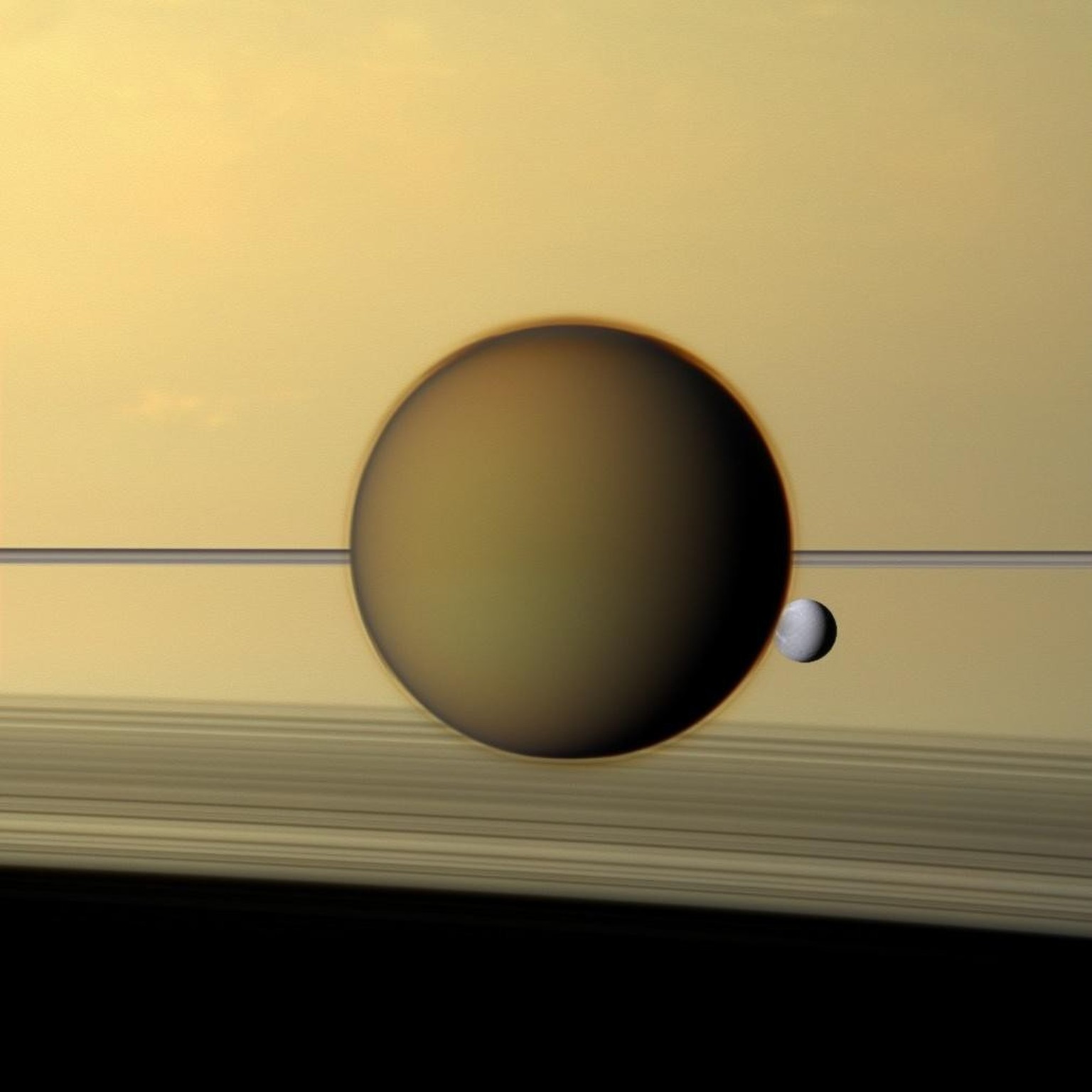 Saturn's third-largest moon Dione can be seen through the haze of its largest moon, Titan, in this view of the two posing before the planet and its rings from NASA's Cassini spacecraft relea ...