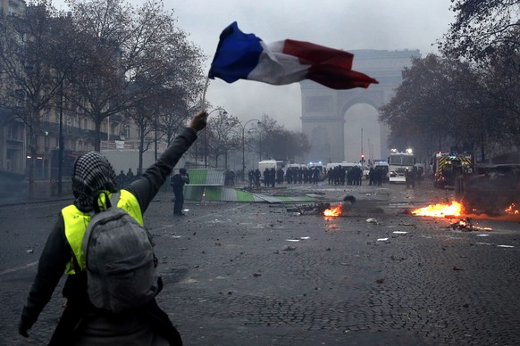 epa07202682 A protester wearing a yellow vest (gilets jaunes) waves a French flag during clashes with riot police near the Arc de Triomphe as part of a demonstration over high fuel prices on the Champ ...