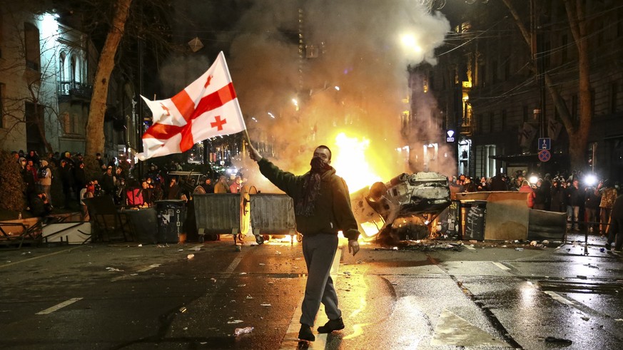 A man waves a Georgian national flag in front of a burning barricade as other protesters stand behind not far from the Georgian parliament building in Tbilisi, Georgia, Thursday, March 9, 2023. Police ...