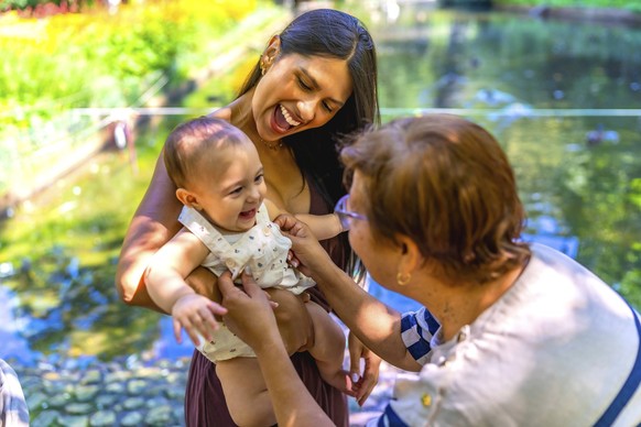 Glückliche lateinamerikanische Familie genießt einen sonnigen Tag in einem Park, die Mutter hält ihr Baby, während die Großmutter mit ihr spielt Happy latin family enjoying a sunny day in a park, moth ...