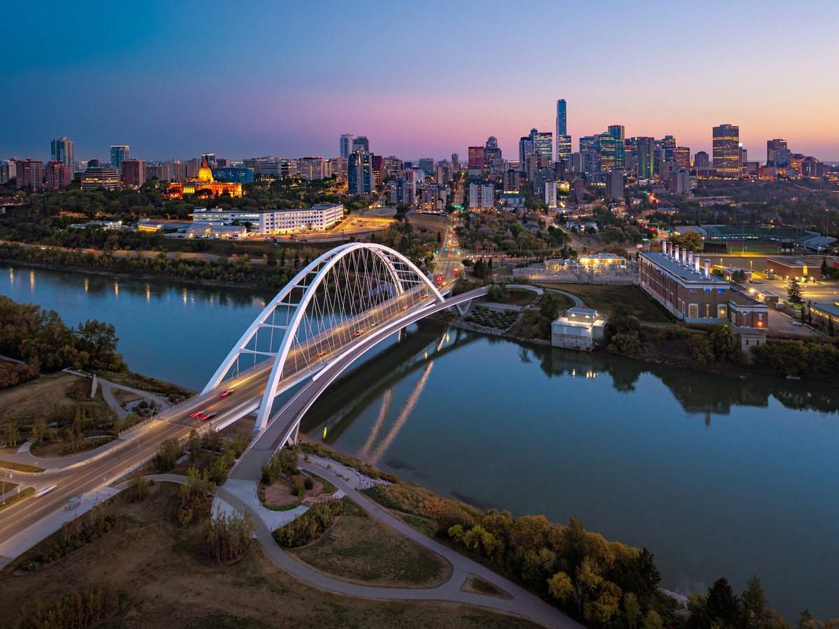 Aerial view of the Walterdale Bridge and downtown Edmonton skyline at dusk, with the North Saskatchewan River reflecting the warm city lights. The modern bridge and illuminated high-rises represent th ...
