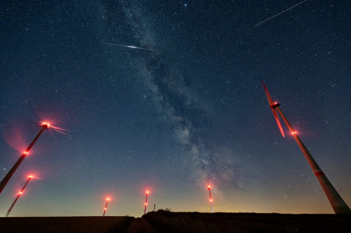 11.08.2023, Hessen, Heppenheim: Die Milchstraße leuchtet über einem Windpark in der Nähe von Schleiden in der Eifel. Die Leuchtspur eines Flugzeuges und die Reflektion eines Satelliten kreuzen das Bil ...