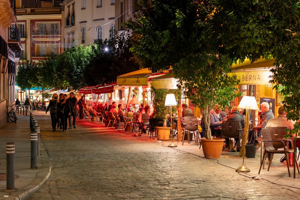Seville, Spain - September 26 2022: A colorful orange tree lined cobblestone road of sidewalk cafes illuminated in the evening in the Barrio Santa Cruz historic district of Seville, Spain.