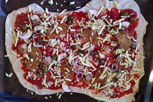 Bereit für den Ofen: die Pizza. Hier mit Champignons, Kapern, Tomatensugo, Schalotten und veganem Salami und veganem Käse belegt.