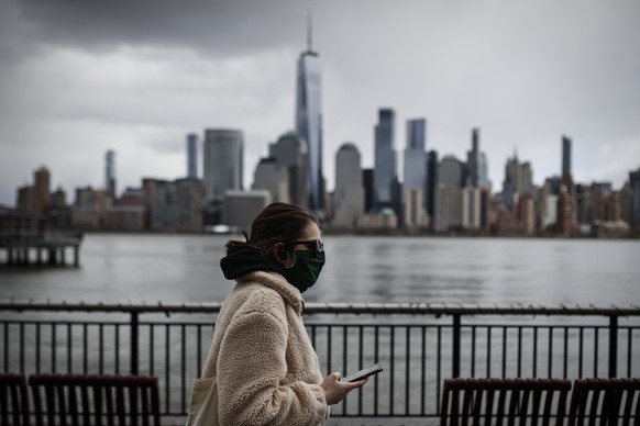 FILE- In this April 10, 2020 file photo a woman wearing a face mask sure to CVID-19 concerns walks along the Jersey City waterfront with the New York City skyline in the background. New York City may  ...