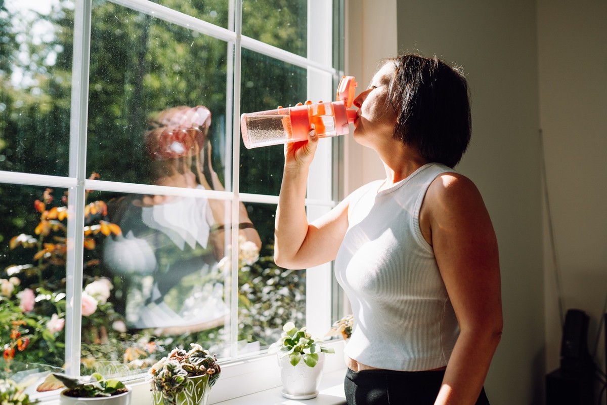 Replenishment of water balance. Mature woman in sportswear drink water after home exercising staying against large window