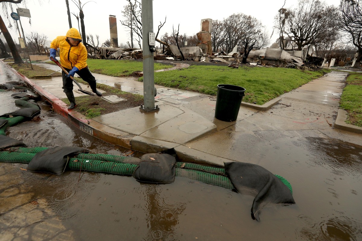 PACIFIC PALISADES, CA - FEBRUARY 13, 2025 - - Pacific Palisades resident Nancy Belanger, 65, clears a clogged drain in front of a fire ravaged home along Sunset Blvd. at Drummond Street as rain falls  ...