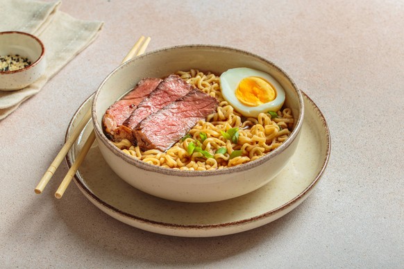 Ramen oder asiatischen Stil Nudelsuppe mit Rindfleisch, Ei und gr�nen Zwiebeln in der Sch�ssel auf hellbeige Hintergrund Bowl filled with ramen noodles, tender sliced beef, boiled egg and fresh green  ...