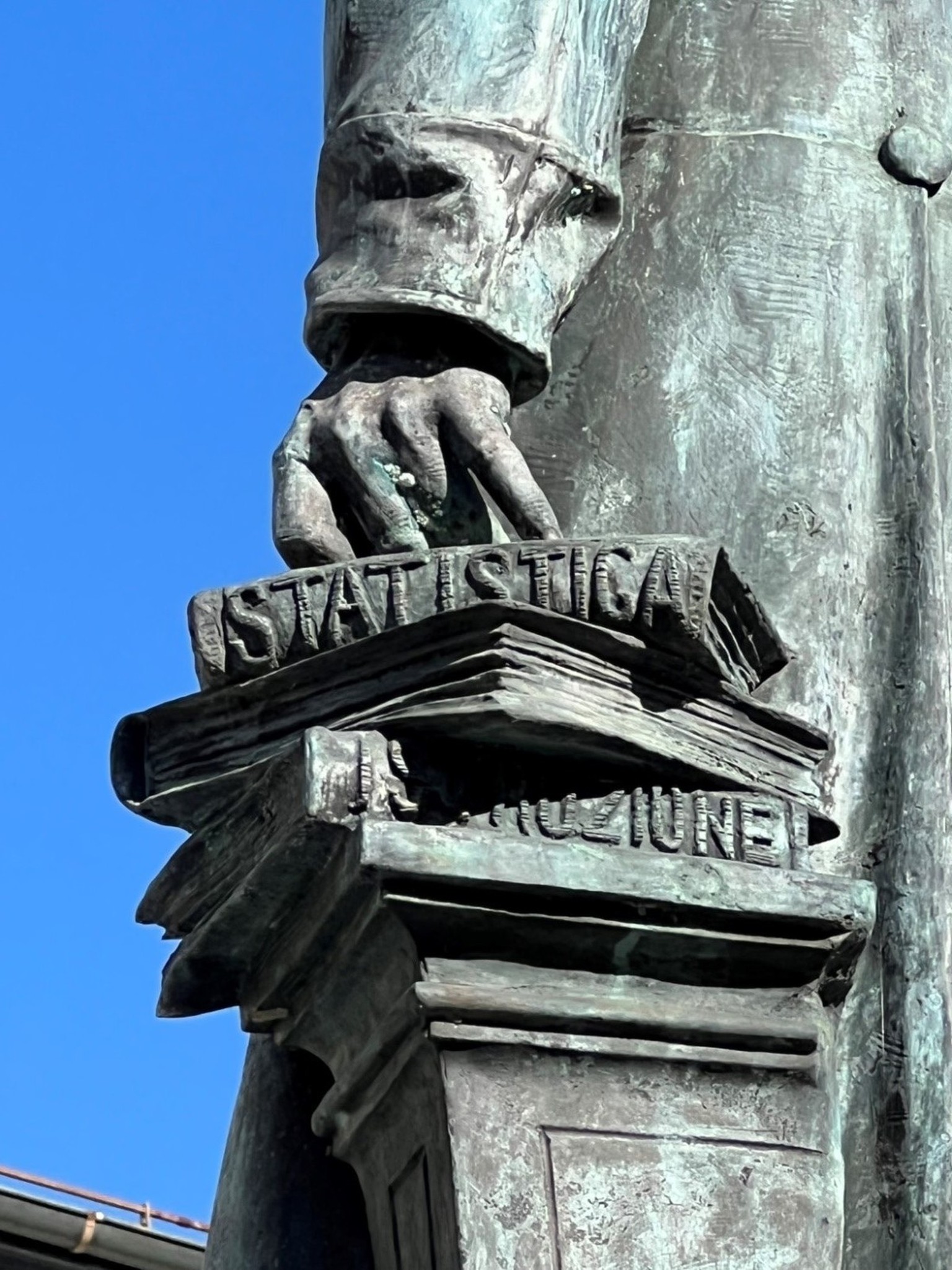 In der Rechten ein Manuskript, die Linke gestützt auf drei Bücher, die sein Wirken verkörpern: STATISTICA, [STORIA], ISTRUZIONE. Denkmal für Stefano Franscini in Faido (Detail).
