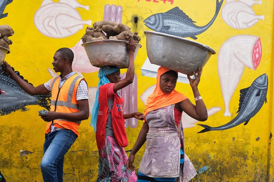 This photo captures a captivating scene of two women carrying aluminum baskets on their heads on their way to the market and walking past a wall covered in vibrant paintings.  One of the women is about...