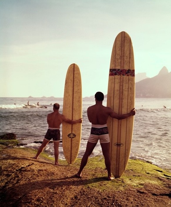 View of two male surfers stand facing the ocean, holding their surfboards vertically beside them, while watching other surfers on the water, California, 1965. (Photo by Tom Kelley/Getty Images)