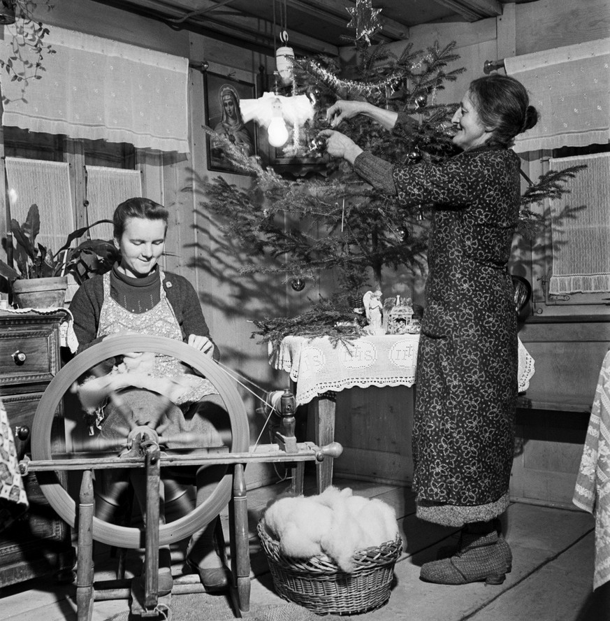 Eine juengere Frau sitzt am Spinnrad, waehrend eine aeltere Frau den Christbaum schmueckt. Weihnachtsvorbereitungen, aufgenommen in Stierva, Kanton Graubuenden, am 24. Dezember 1941. (KEYSTONE/PHOTOPR ...