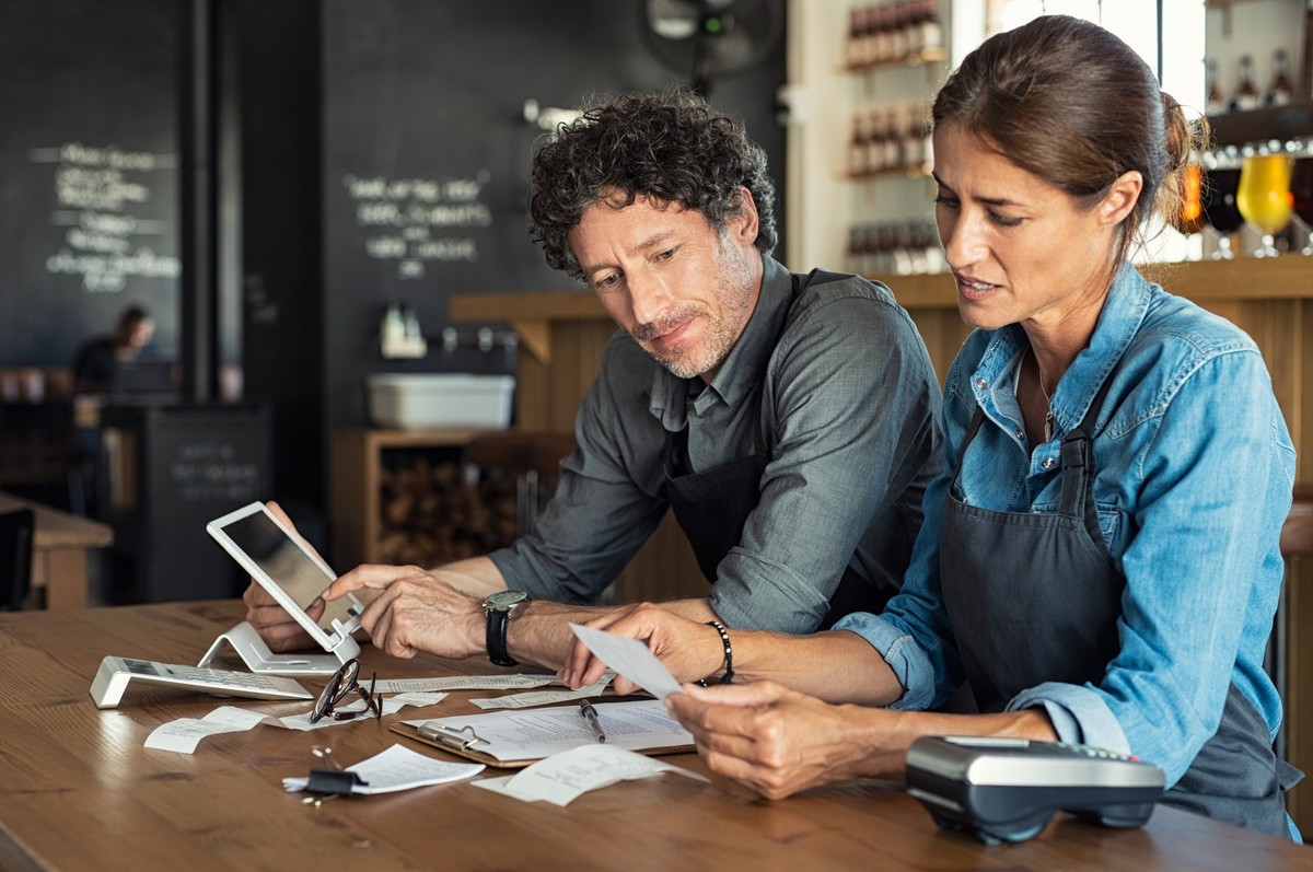 Man and woman sitting in cafeteria discussing finance for the month. Stressed couple looking at bills sitting in restaurant wearing uniform apron. Café staff sitting together looking at expenses and b ...