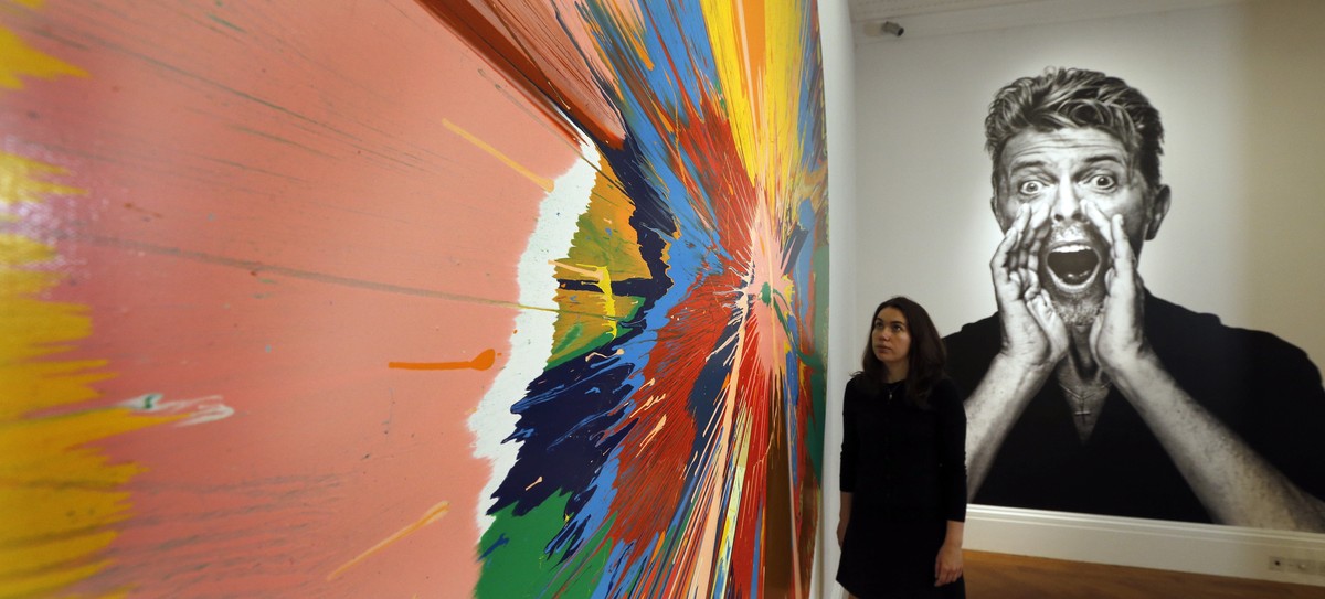 A woman looks at a painting by Damien Hirst in front of a portrait of David Bowie at Sotheby's in London, Wednesday, July 20, 2016. Sotheby's employees lift the painting "Air Power&quot ...
