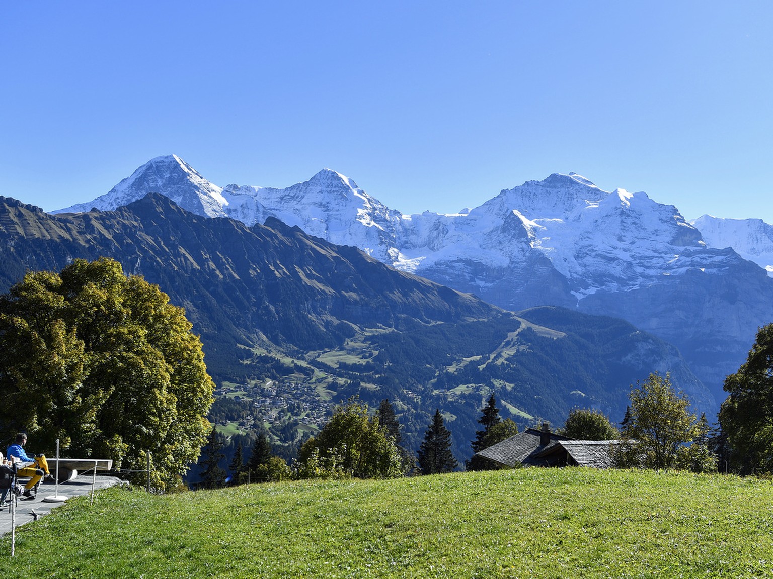 Personen geniessen auf einer Terasse das schoene Herbstwetter mit Blick auf das Dreigestirn Eiger, Moench und Jungfrau, von links, am Sonntag, 16. Oktober, in Sulwald ob dem Lauterbrunnental. (KEYSTON ...