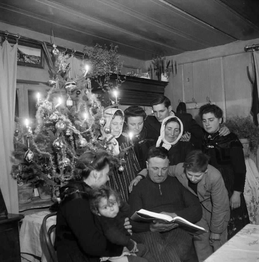 Frauen, Maenner und Kinder versammeln sich in der Wohnstube neben dem geschmueckten und mit Weihnachtskerzen illuminierten Christbaum. Der aeltere Mann liest aus der Bibel vor. Aufmerksam verfolgt von ...