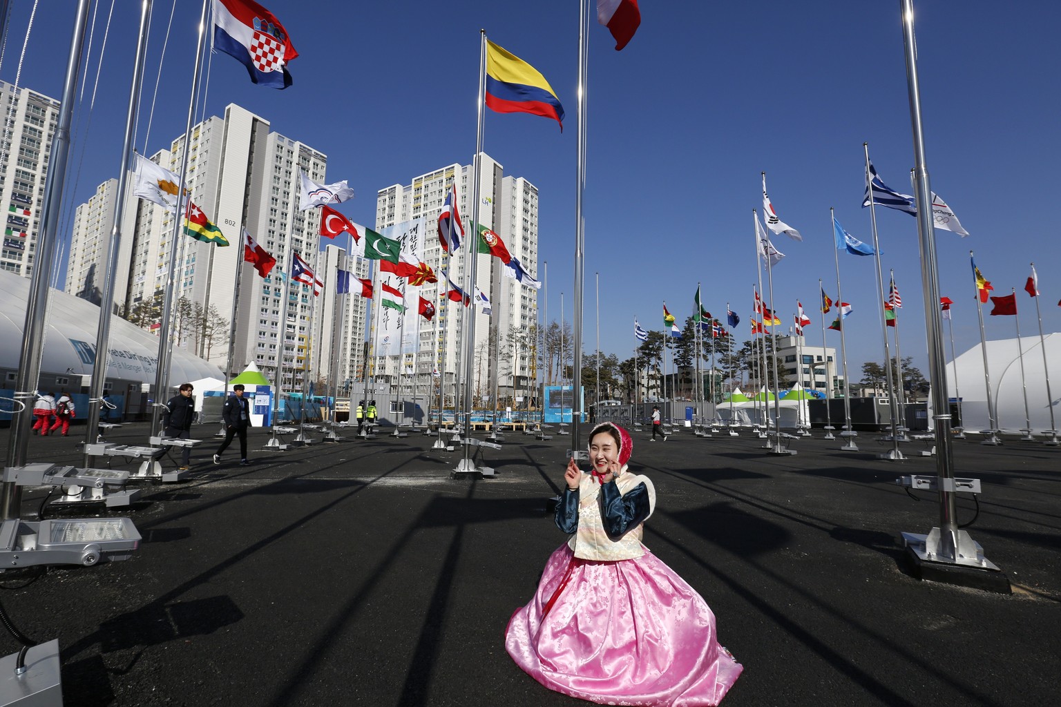 epa06495230 A girl dressed in traditional clothes poses for a photo taken by a friend in front of the flags of competing nations and the Olympic athletes accommodation village (high rises in backgroun ...