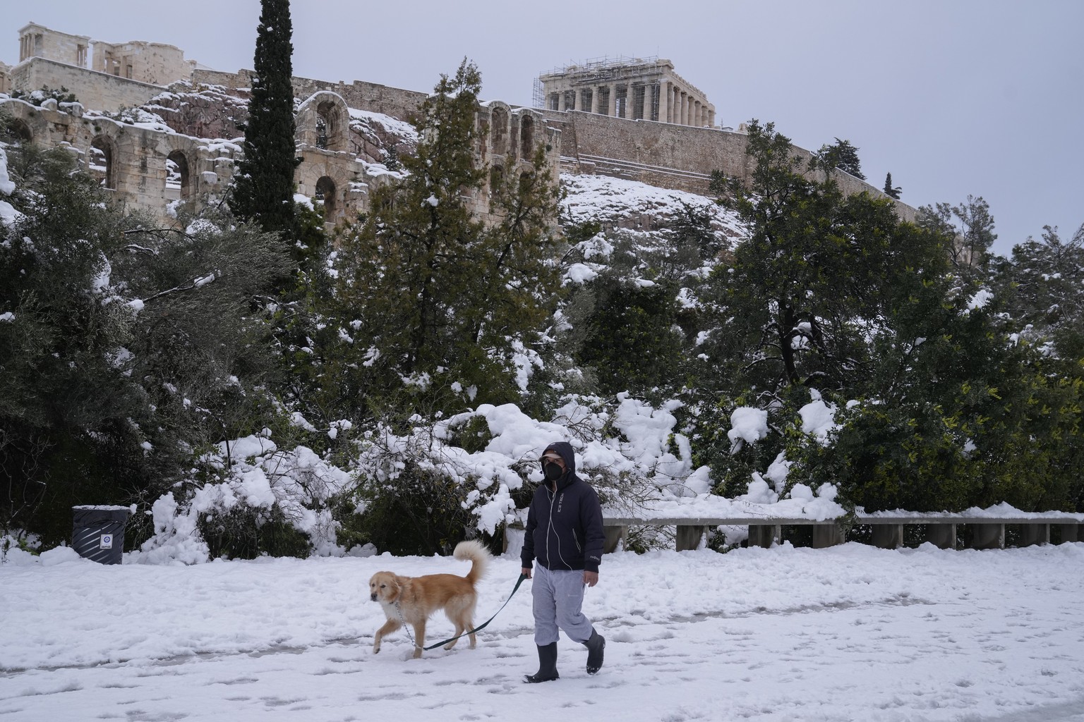 A pedestrian walks with his dog on the snow in front of the ancient Acropolis hill, in Athens, on Tuesday, Jan. 25, 2022. A snowstorm of rare severity disrupted road and air traffic Monday in the Gree ...
