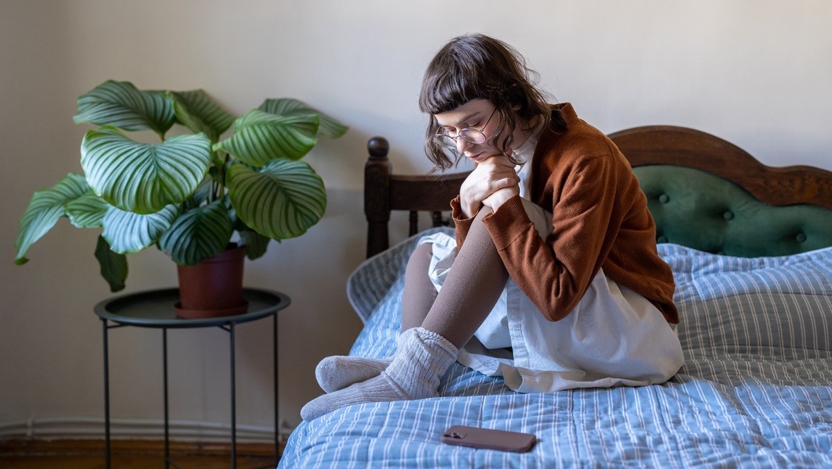 Sad depressed teenage girl in eyeglasses sitting on bed waits for call from boyfriend in difficult relations. No answer phone message. Loneliness without cellphone sms. Network internet addiction xkwx ...
