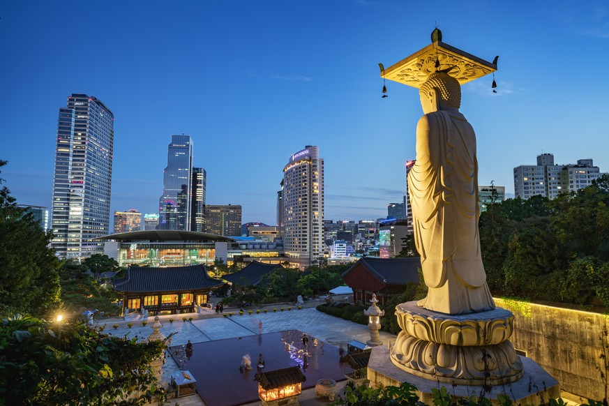 Famous Bongeunsa Temple Buddha Statue at Night - Twilight. Modern Skyscrspers of downtoen Seoul illuminated in the background. Half aerial view from the jungle behind the temple.