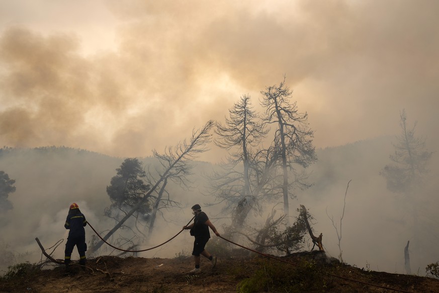 A firefighter tries to extinguish the flames as a local resident holds a water hose during a wildfire at Ellinika village on Evia island, about 176 kilometers (110 miles) north of Athens, Greece, Mond ...