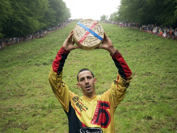 Chris Anderson, 30, holds up his prize after breaking the all-time record for the most cheeses won in the death-defying Cheese Rolling Race, in Brockworth, Gloucestershire, England, Monday May 28, 201 ...