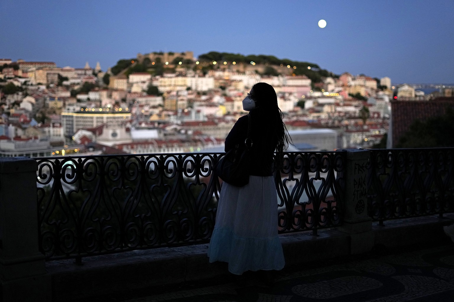 A woman wearing a face mask looks at downtown Lisbon, Portugal, from a viewpoint in Bairro Alto, or high quarter, as the moon rises after sunset, Sunday, Sept. 19, 2021. (AP Photo/Armando Franca)