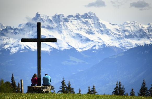 People take a break under a cross in a field and enjoy a spring weather day in front of the Swiss Alps mountains (Les Dents du Midi), in Les Pleiades Blonay above Montreux, Switzerland, Sunday, May 23 ...